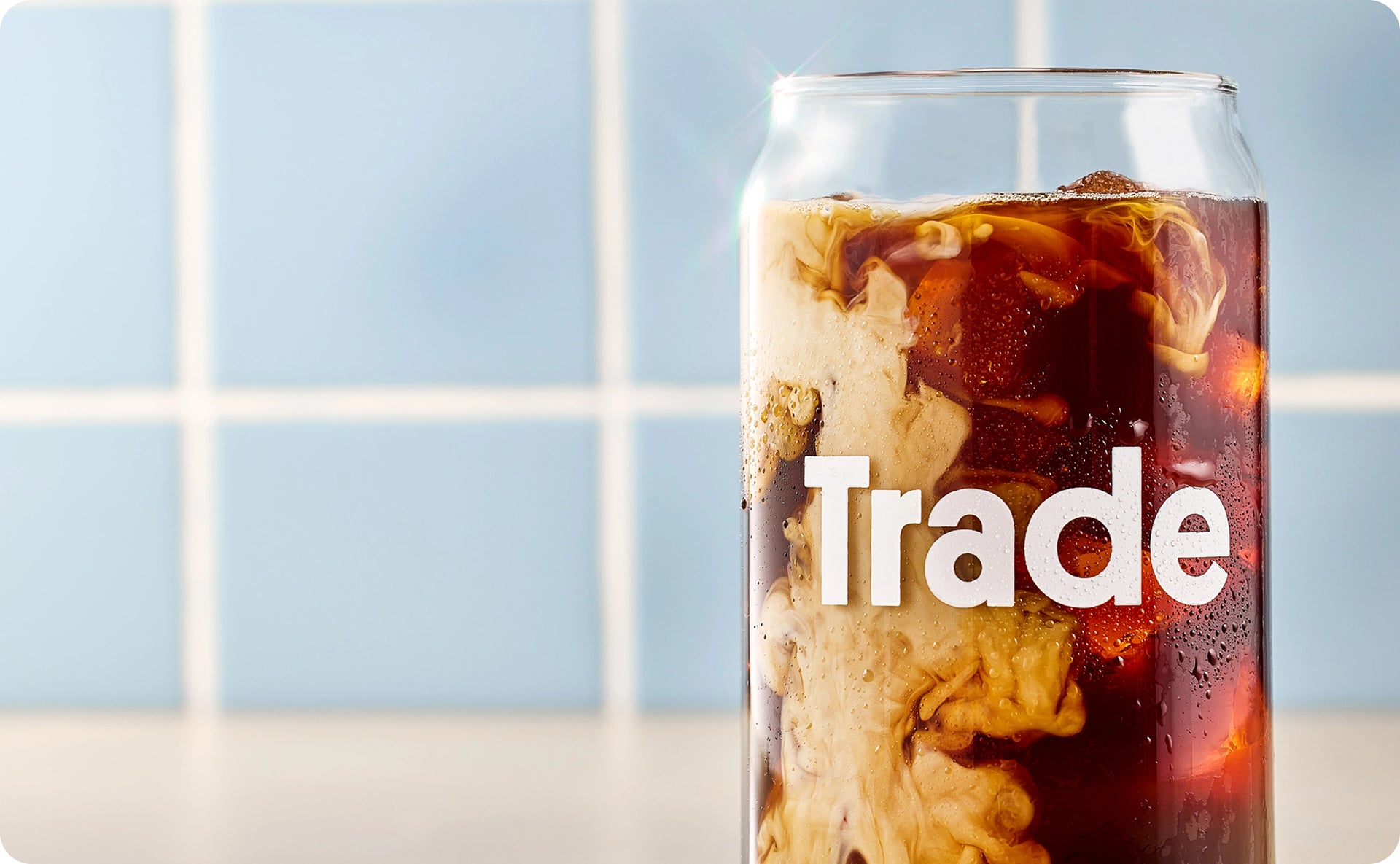 A tall glass can filled with iced coffee and cream swirling through it, with the Trade logo in white text, against a light blue tiled background.