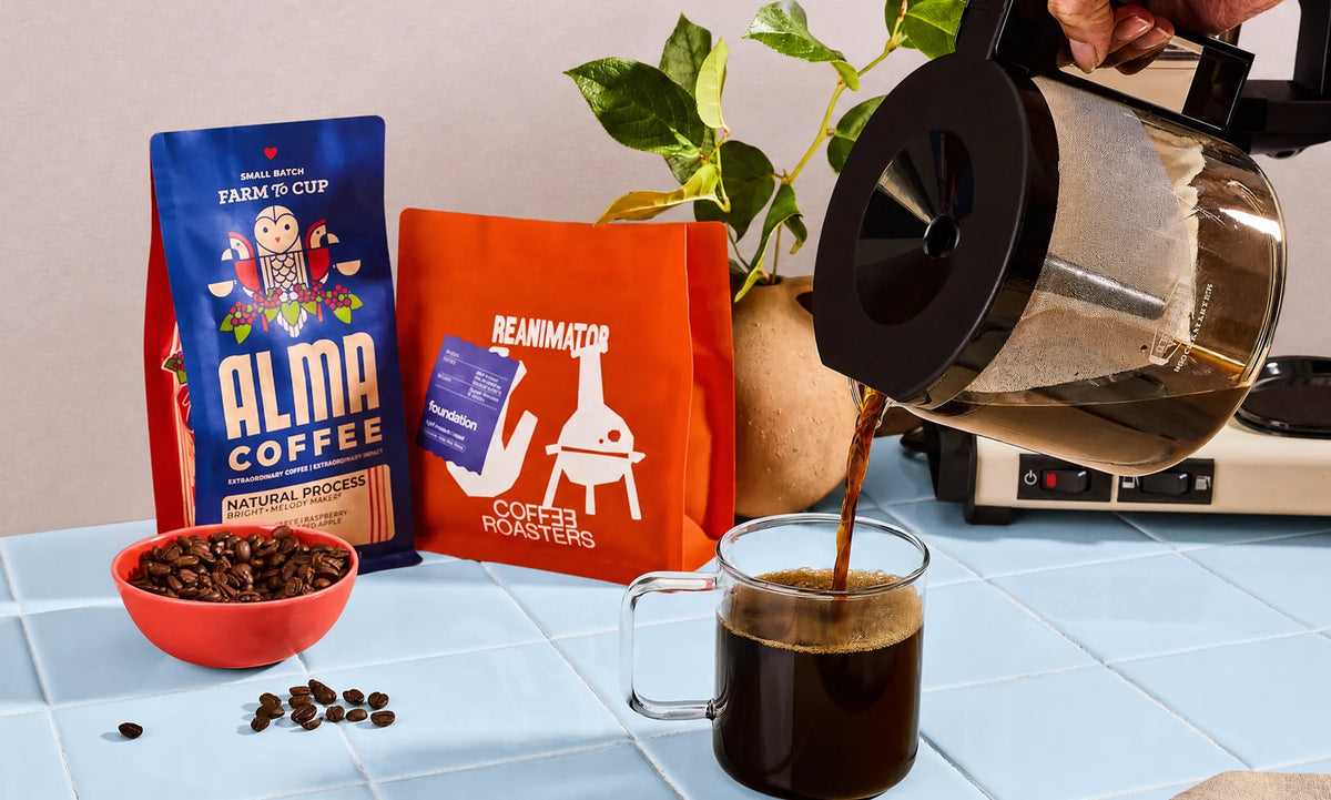 Coffee pouring from a glass carafe into a mug, beside bags of Alma Coffee and Reanimator Coffee Roasters