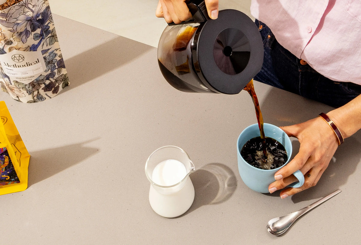 Coffee pouring from a glass carafe into a light blue mug, with a small glass pitcher of milk and a bag of Methodical Oscuro coffee nearby.