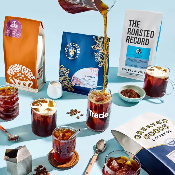 Coffee beans being poured into a glass with coffee bags and cups in the background on a light blue surface.