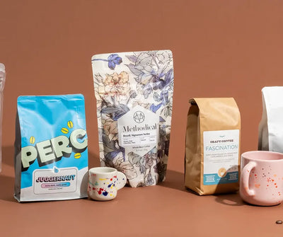 Assortment of colorful coffee bags and speckled ceramic mugs arranged on a terracotta background