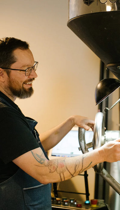 Bearded roaster in glasses smiling while operating a commercial coffee roasting machine